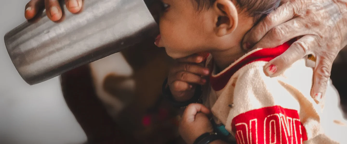 Baby supported by an adult while sipping from a stainless steel cup indoors