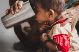 Baby supported by an adult while sipping from a stainless steel cup indoors