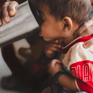 Baby supported by an adult while sipping from a stainless steel cup indoors