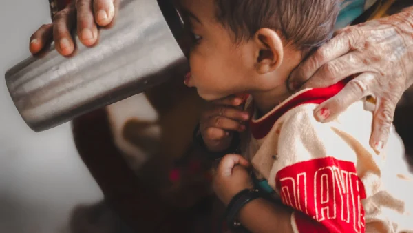 Baby supported by an adult while sipping from a stainless steel cup indoors