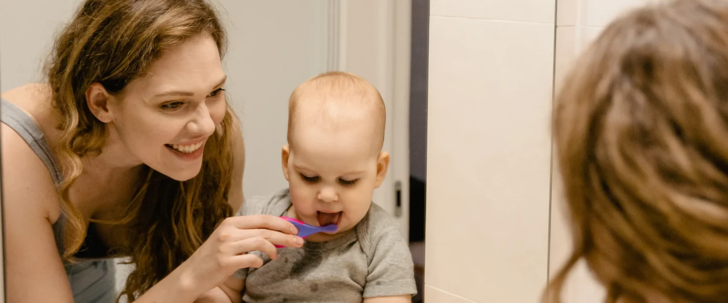 Mother helping her baby brush teeth while looking in a bathroom mirror