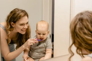 Mother helping her baby brush teeth while looking in a bathroom mirror