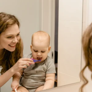 Mother helping her baby brush teeth while looking in a bathroom mirror