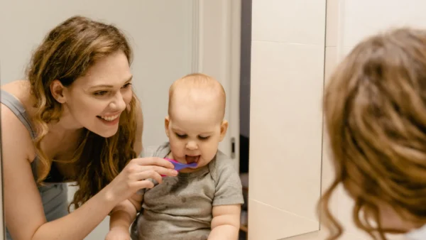 Mother helping her baby brush teeth while looking in a bathroom mirror