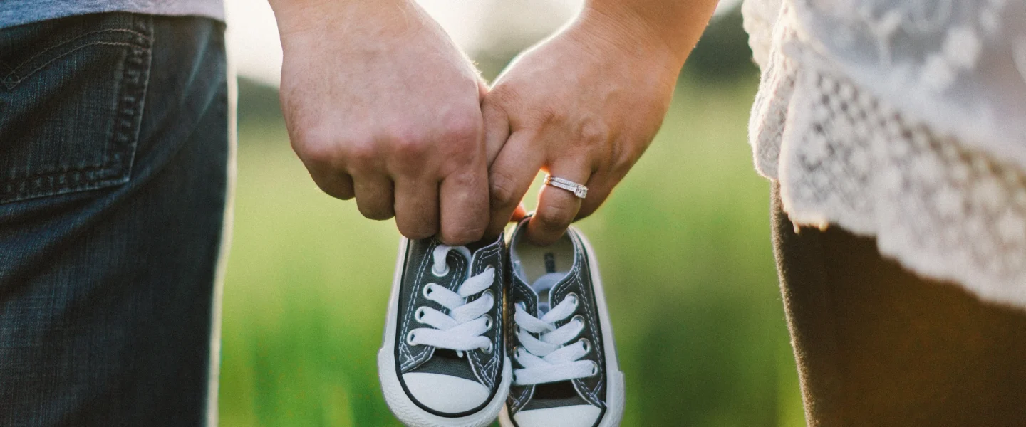 Two adults holding a tiny pair of dark canvas baby sneakers outdoors
